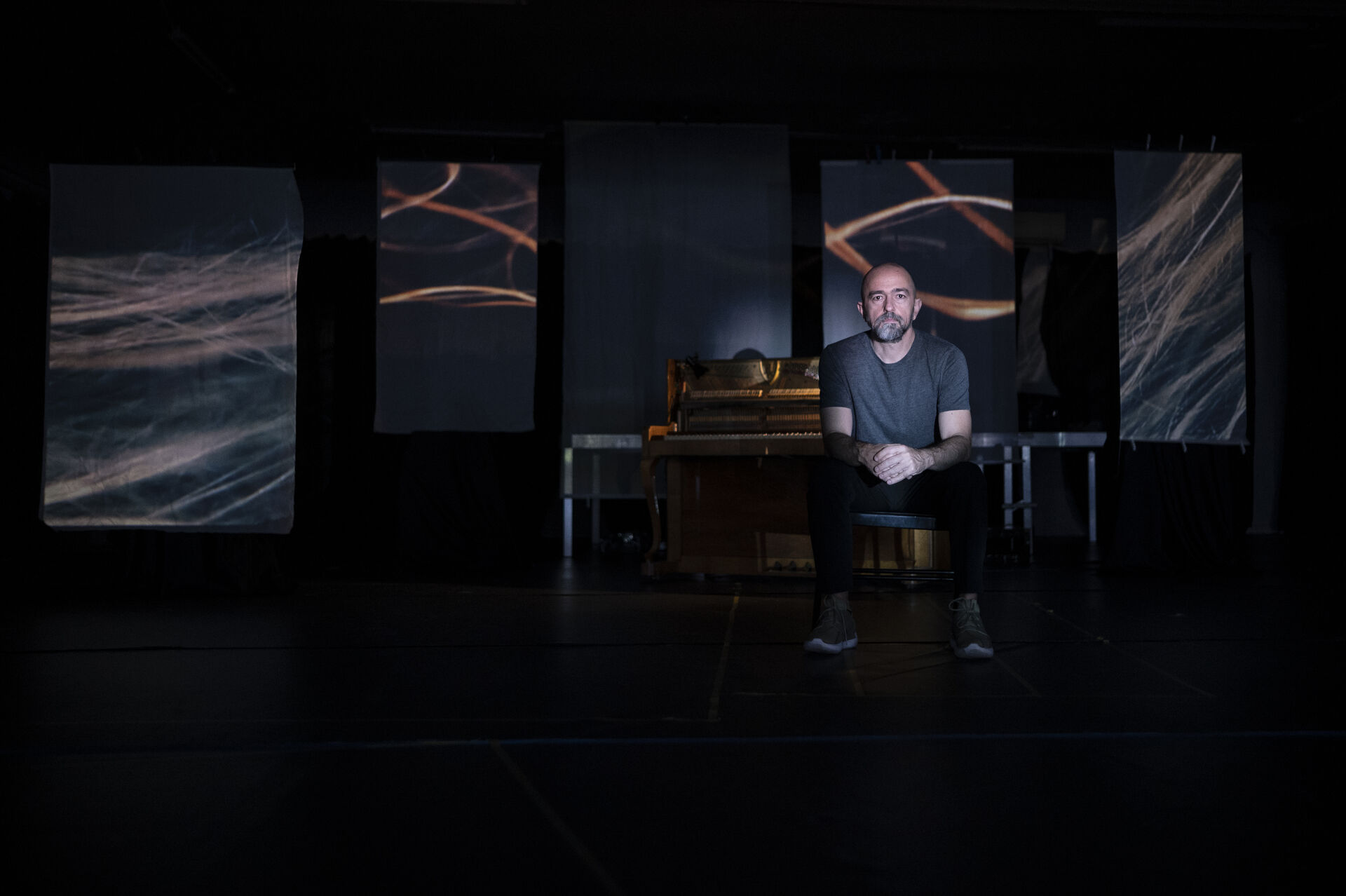 Expanded Piano II: Gasparatos seated by a piano on a dark stage, surrounded by hanging printed banners showing wave-like sound visualisations. Photo by Γιώργος Καλκανίδης.